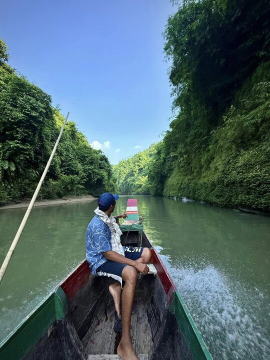 A long boat running up a narrow river walled by forest, Bandarban.