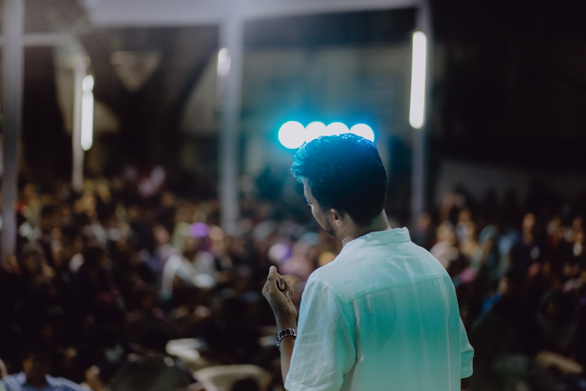 Seen from behind in a white shirt, facing a large seated crowd, blue lights overhead.