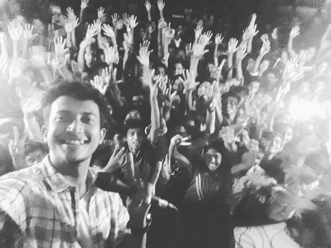 A black-and-white selfie, smiling, with a crowd raising their hands behind.