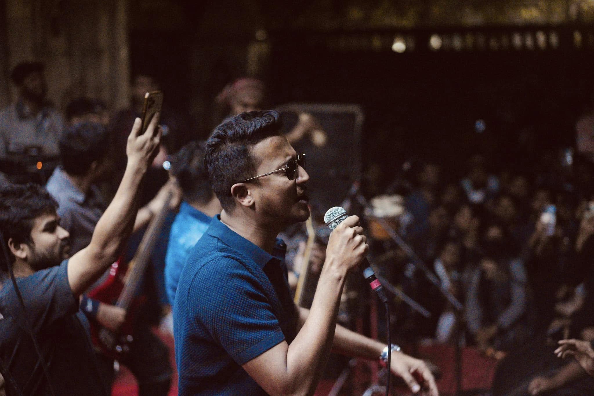 A singer in sunglasses and a navy shirt sings into a microphone while a fan beside him holds up a phone.
