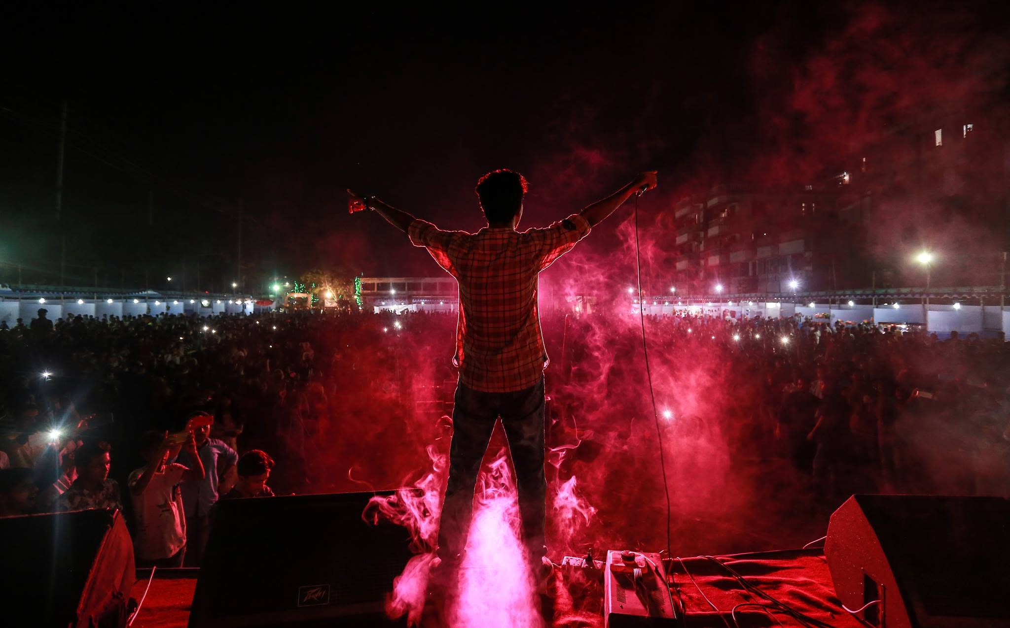 Seen from behind on a stage, arms spread wide towards a large outdoor crowd at night, red smoke rising.