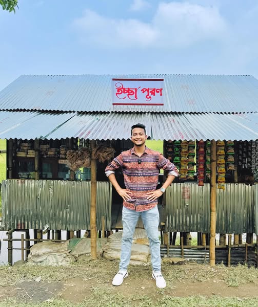 Tasrif in front of a small village shop, stocked, under a hand-painted sign reading ইচ্ছা পুরণ.