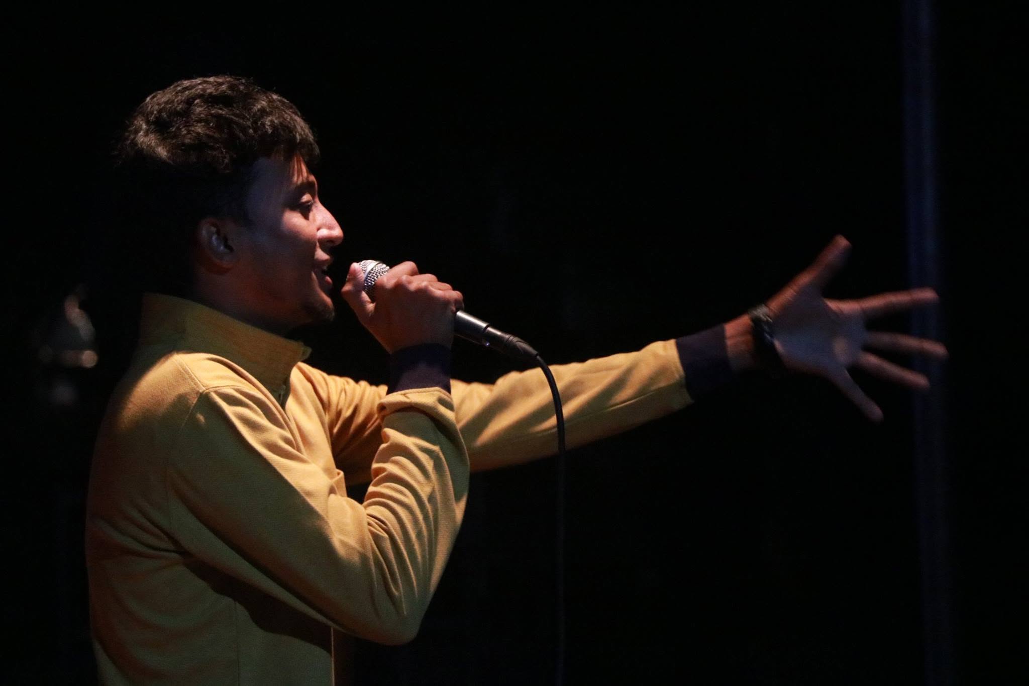 A singer in a mustard shirt sings into a microphone, his other arm stretched out, against a dark background.