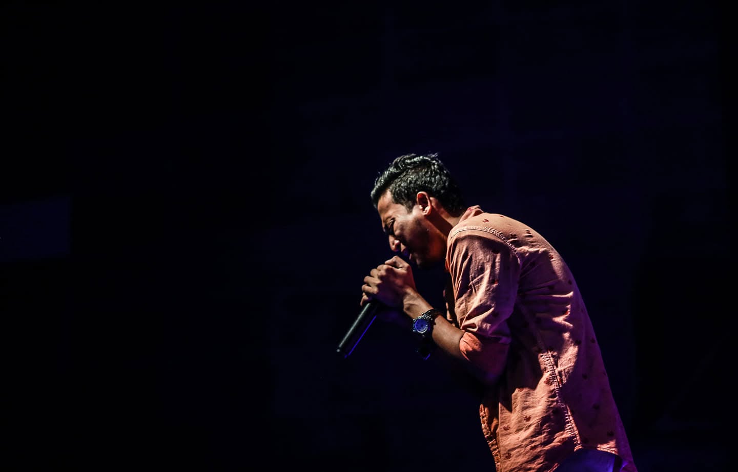A singer bent over a microphone under violet stage light, the stage around him dark.