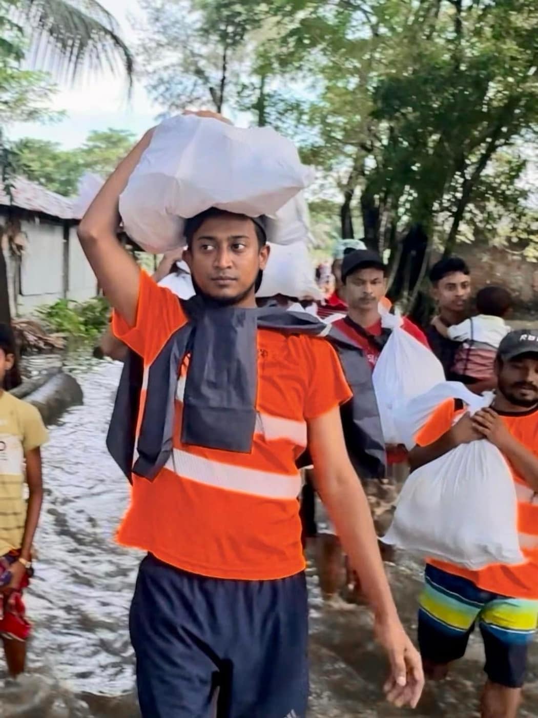 Tasrif wading a flooded lane with a sack on his head, a line of people in high-visibility vests behind him.