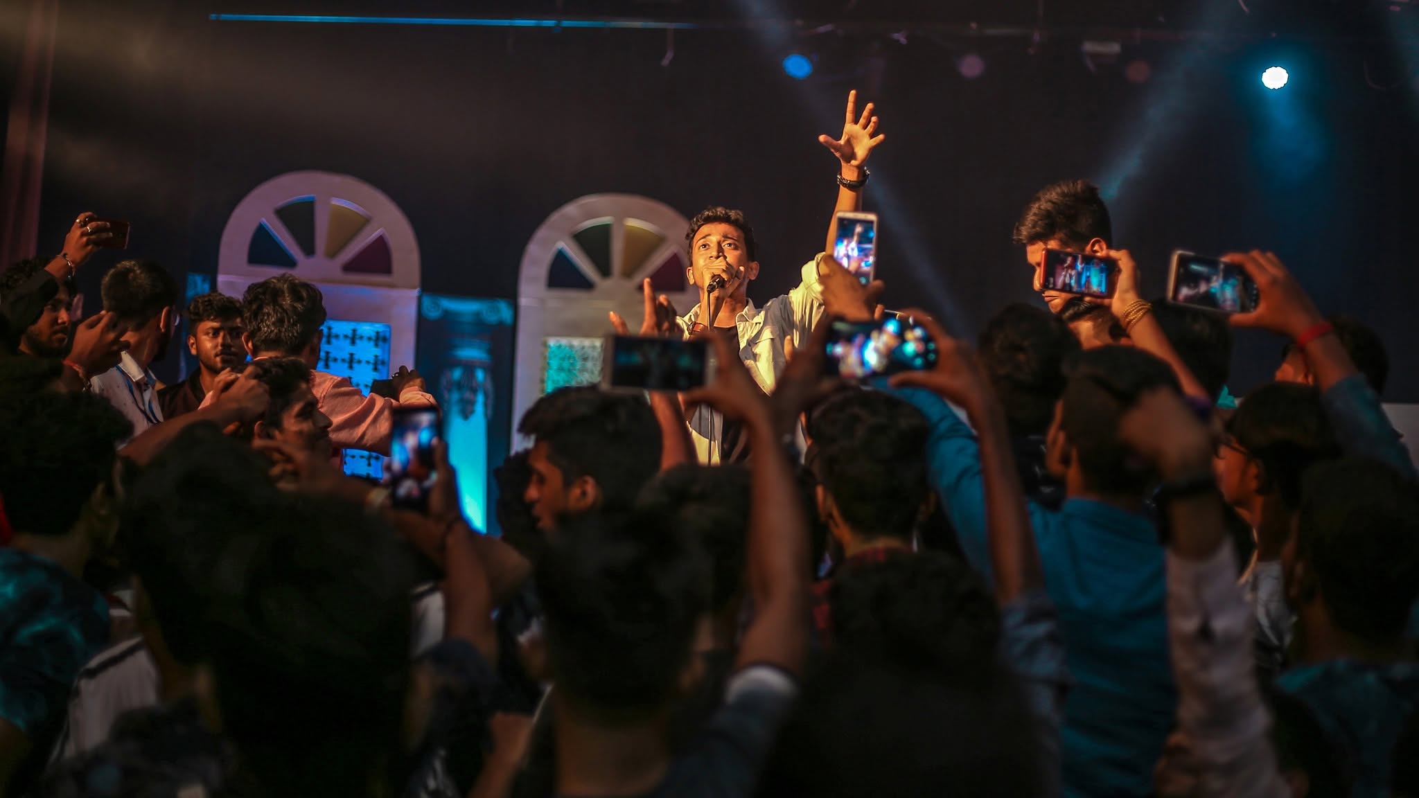 A singer on a stage between two arched windows, one arm raised, while the crowd in front films on their phones.