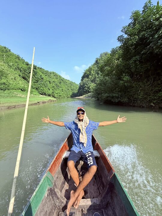 Arms wide at the bow of the boat as it moves upriver, Bandarban.