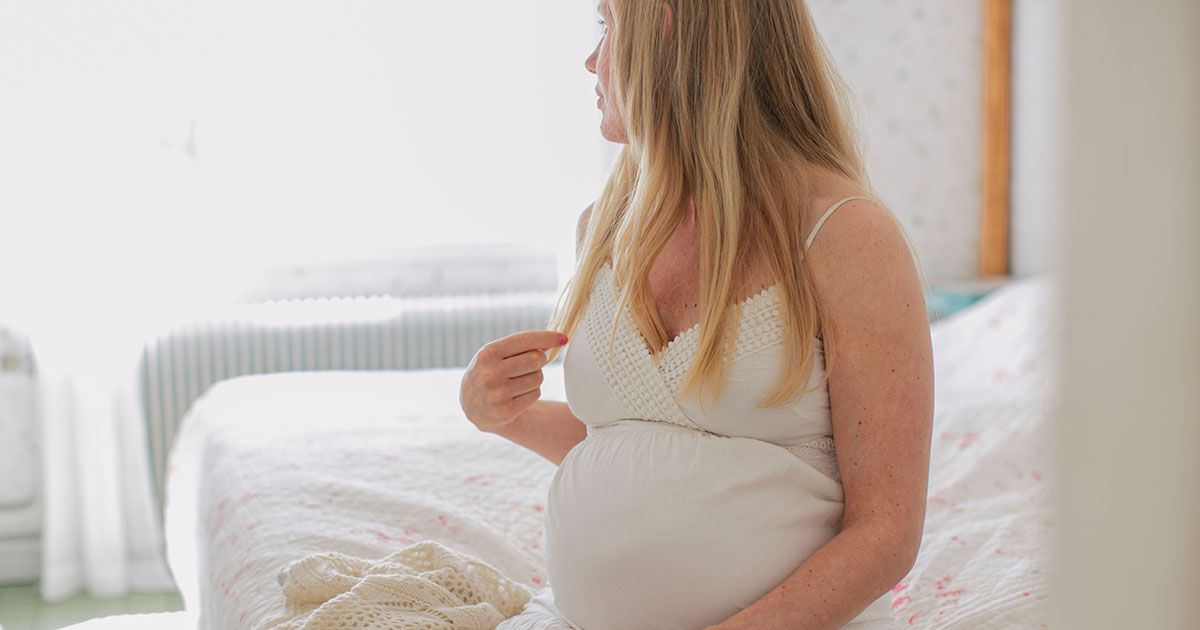 Gravid kvinna, sitter i sängen med vit klänning och tittar bort från kameran.