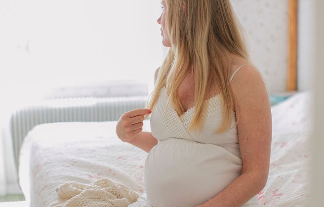 Gravid kvinna, sitter i sängen med vit klänning och tittar bort från kameran.