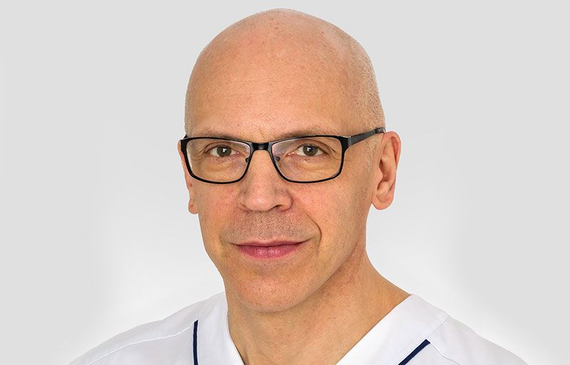 Örjan Friberg ler mot kameran med en grå bakgrund. Han har svarta glasögon på sig.
