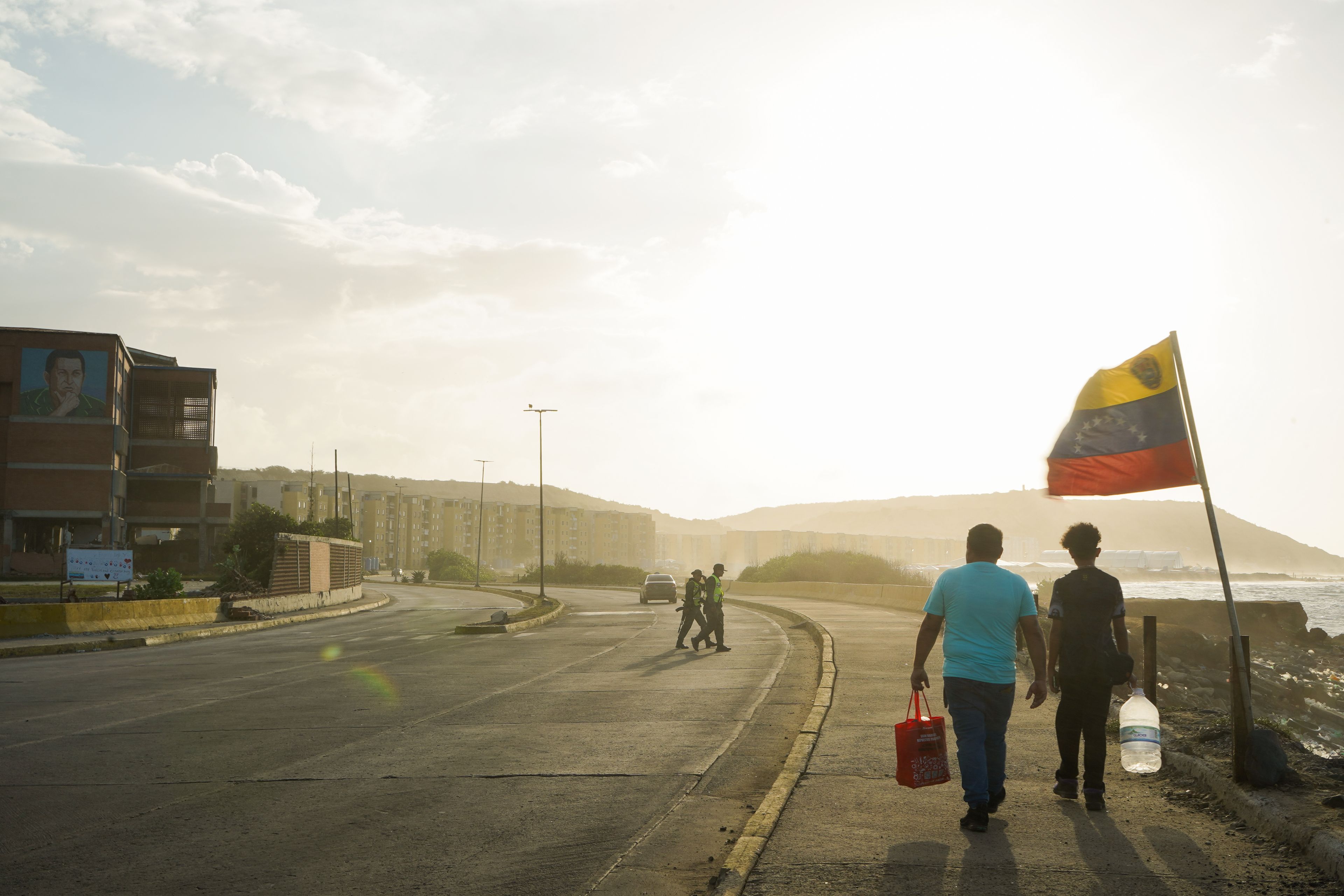 People walk along a road in La Guaira as a Venezuelan flag flies beside the waterfront. Photo: María José Dugarte