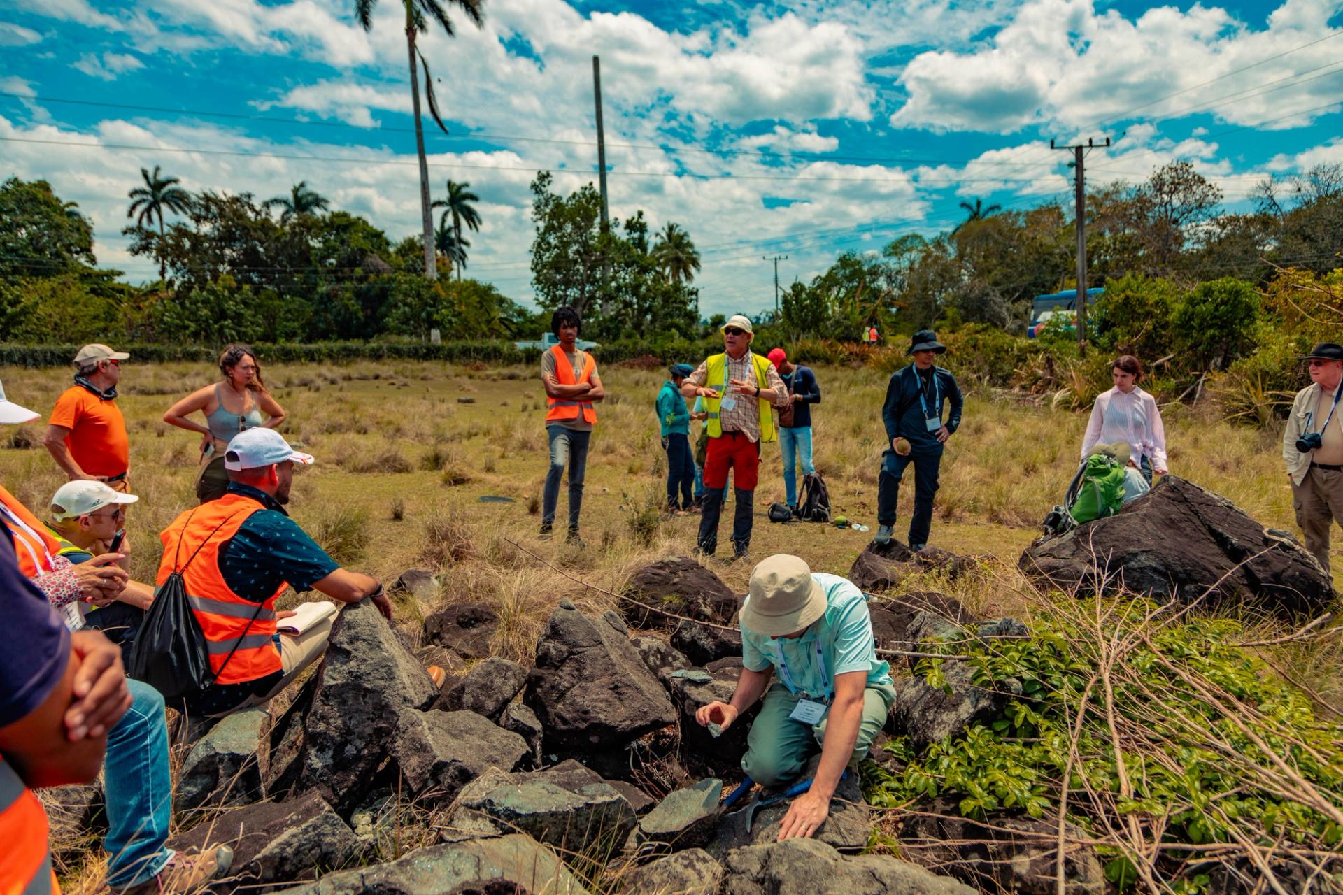 A field group observes a man in a safety vest gesturing, while another crouches in the foreground to inspect a rock among boulders.
