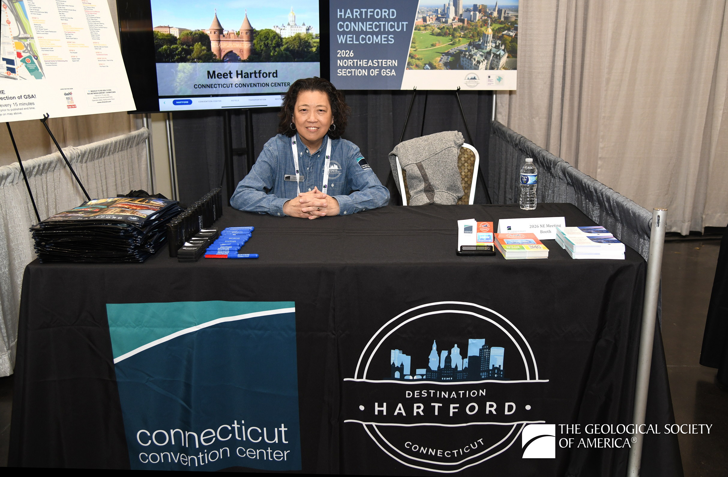 A person sits smiling at a booth with a "Destination Hartford" banner, surrounded by brochures, a water bottle, and promotional materials for Hartford, Connecticut.