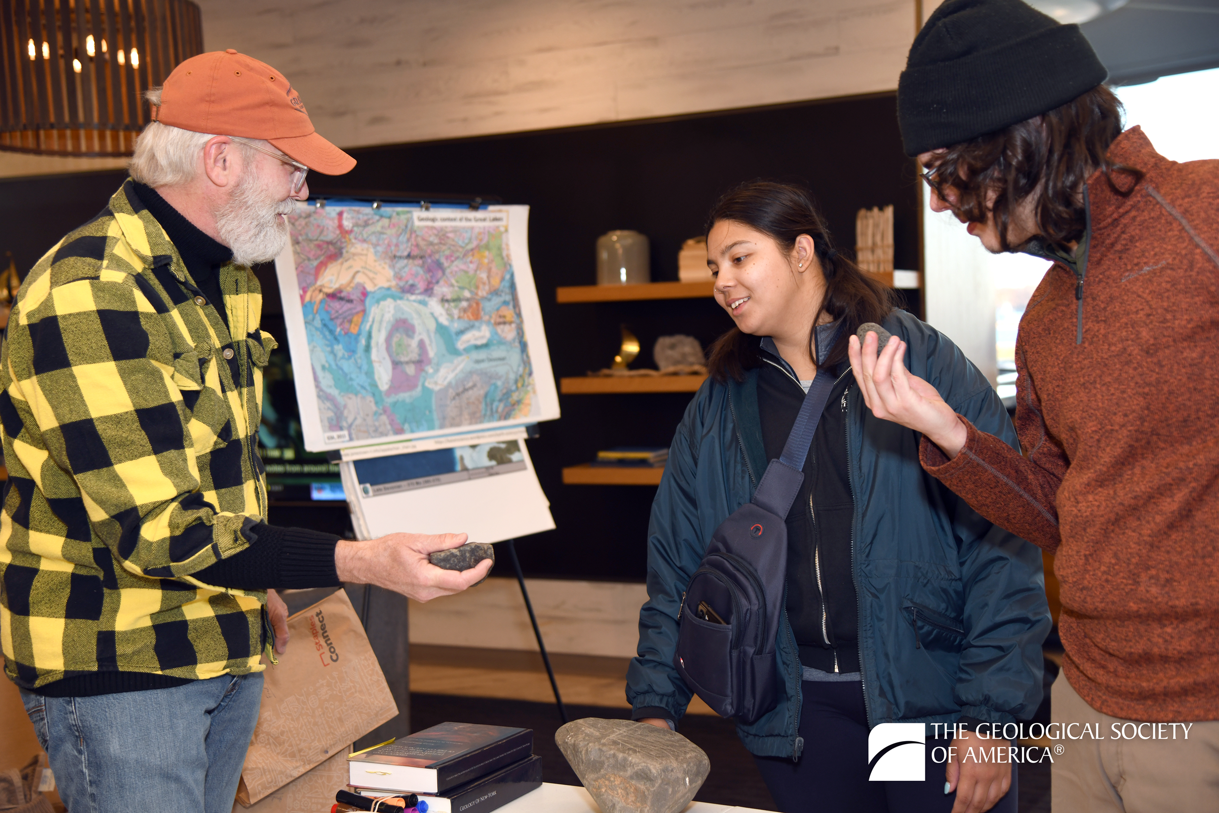 An older man in a yellow plaid shirt shows a rock to two engaged young adults in a cozy room with maps and books, conveying interest in geology.