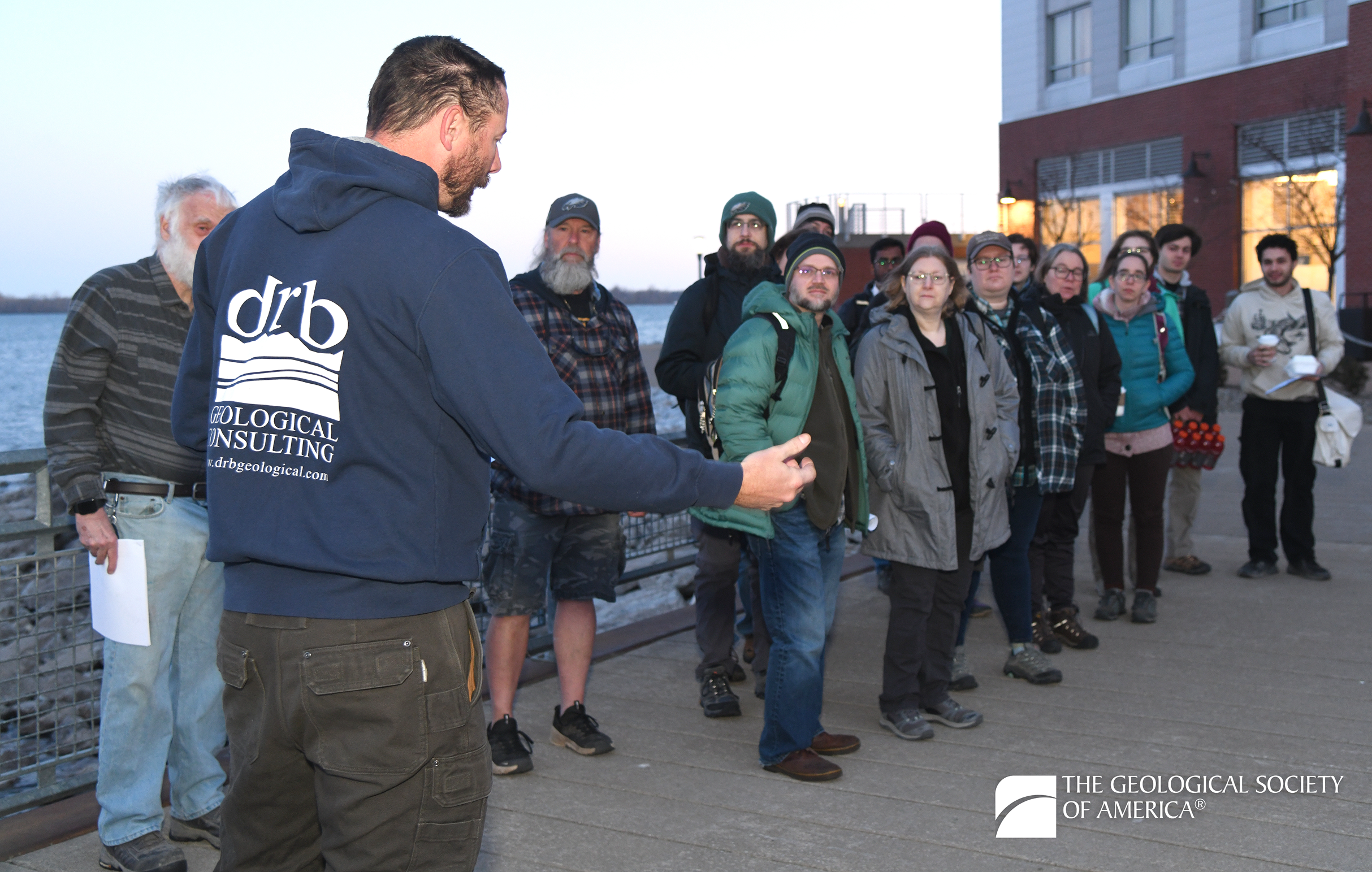 A group of people stand on a boardwalk near the water, listening to a man in a blue hoodie speak. Evening light gives a calm, educational tone.