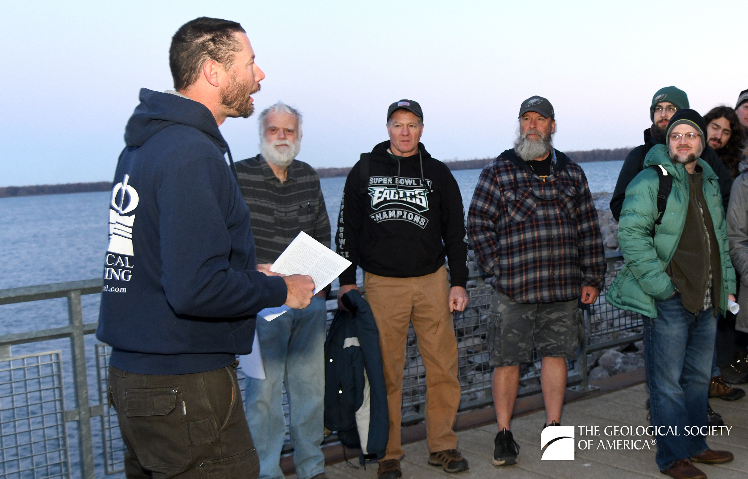 A group of people stands outdoors by a lake at dusk. A man in a hoodie addresses them, holding papers. Atmosphere is informal and attentive. Logo reads "The Geological Society of America."