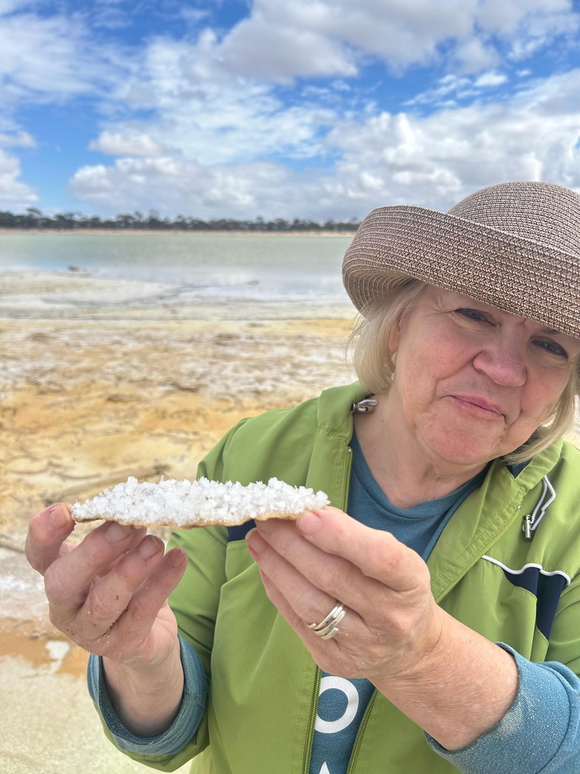 A blonde woman in a straw hat and green shirt layered over a blue shirt holds a fossil on the beach.