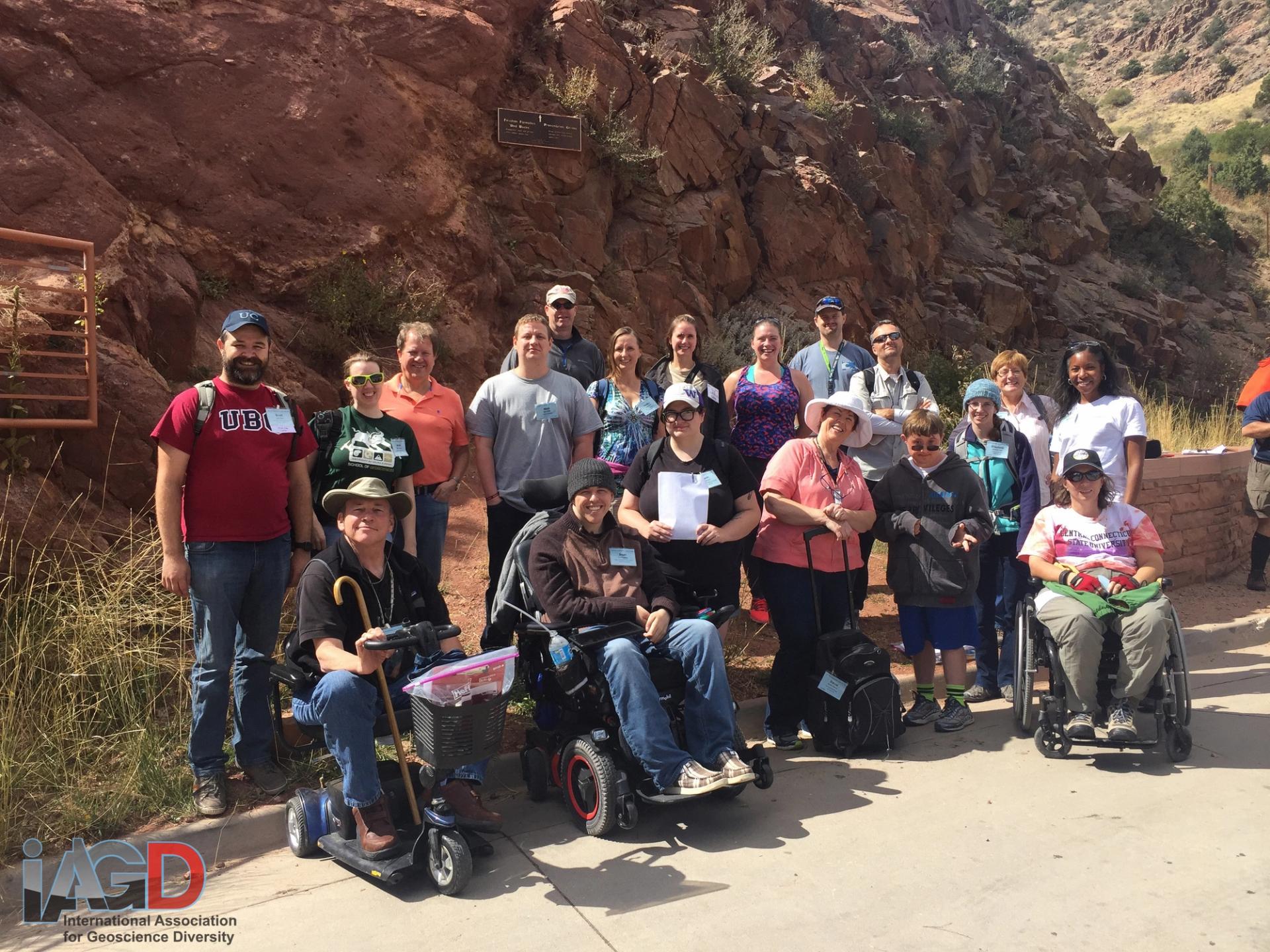 A diverse group of fifteen people, including those in wheelchairs, pose smiling in front of rocky cliffs, suggesting an inclusive outdoor activity.