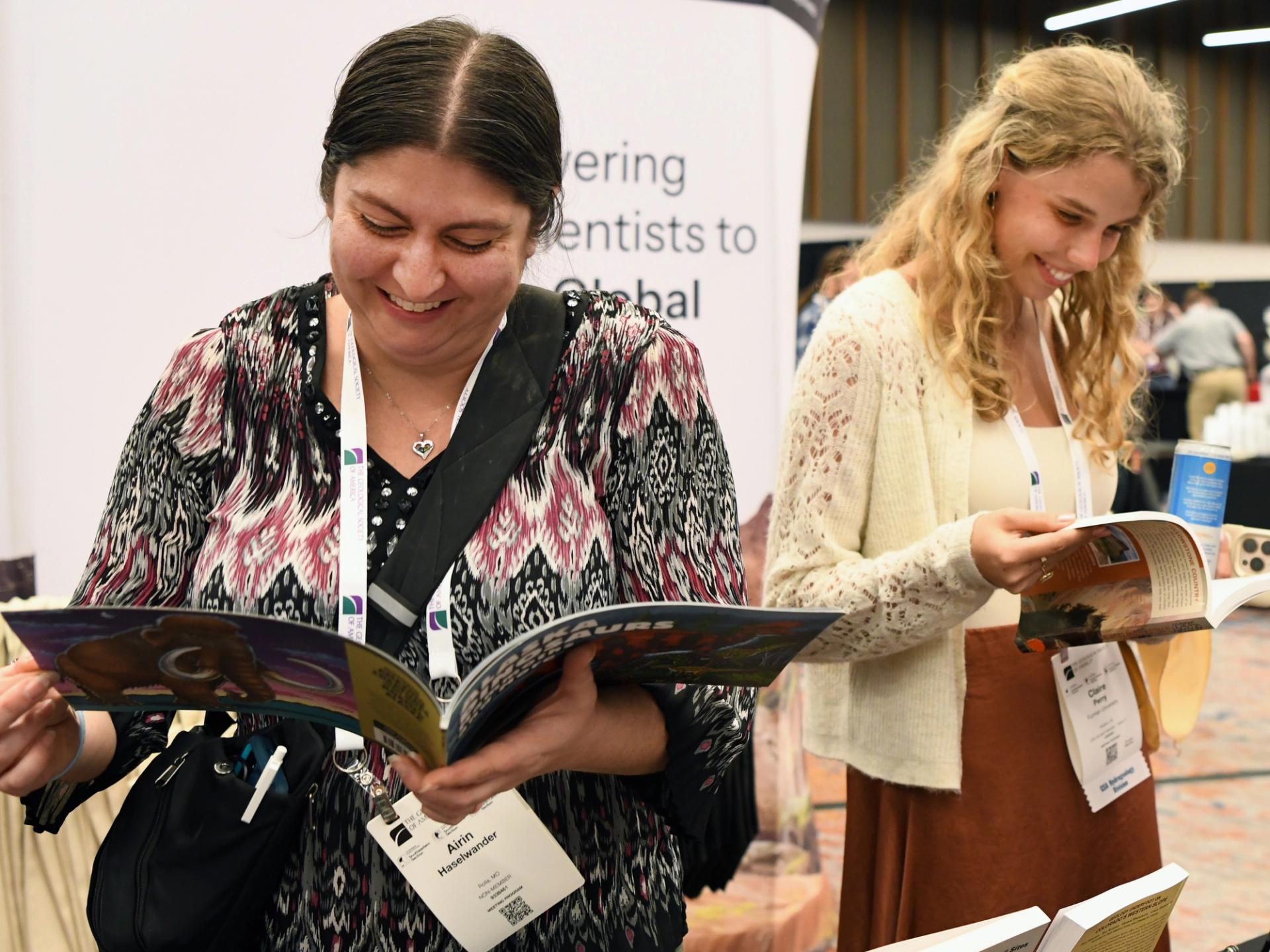 Two women smiling and reading magazines at an indoor event. They wear name tags and appear engaged, creating a warm, lively atmosphere.