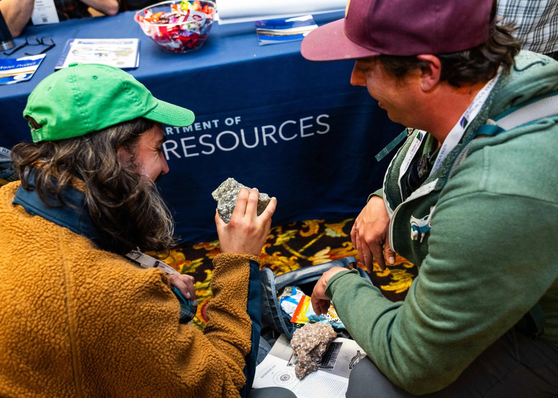 Two people examine rock samples while seated on the floor near an exhibit table at a geoscience event.