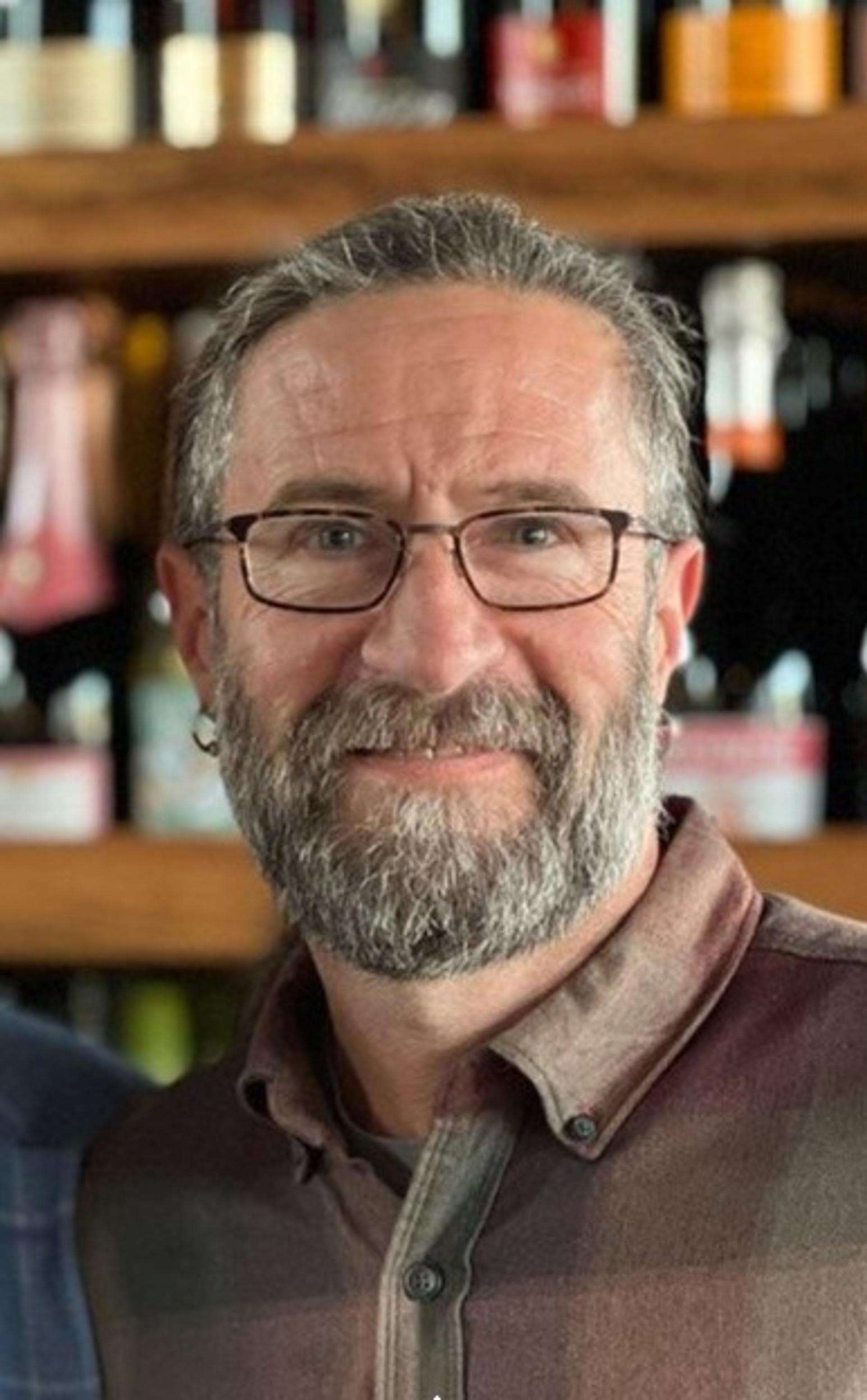 Todd LaMaskin in glasses and a collared, button-up shirt with wooden shelves in the background.