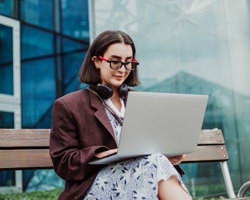 Woman sitting on bench with laptop on her lap and headphones around her neck.