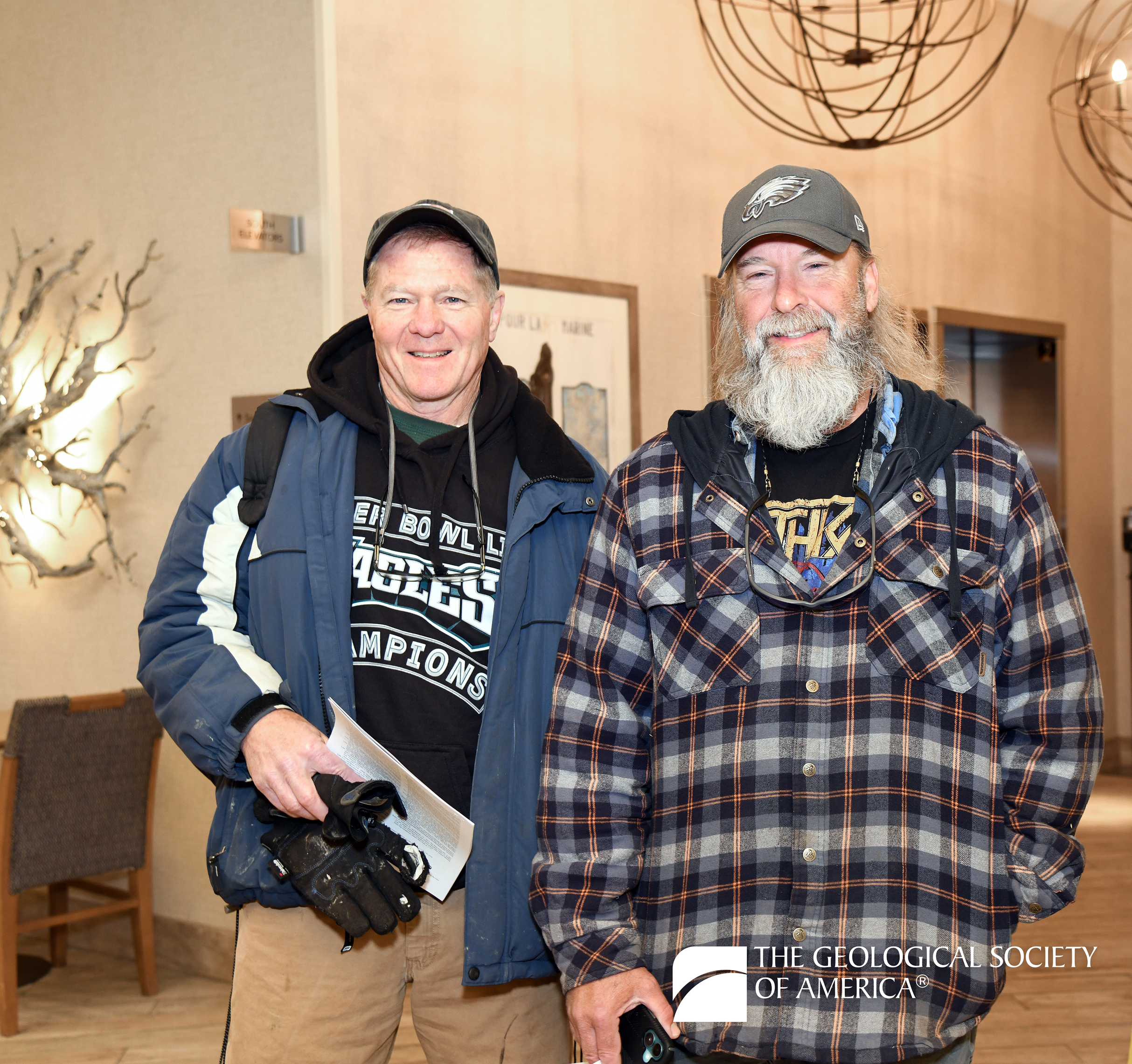 Two smiling men in casual jackets and hats stand indoors, one holding papers and gloves. The setting has modern decor. Geological Society logo present.