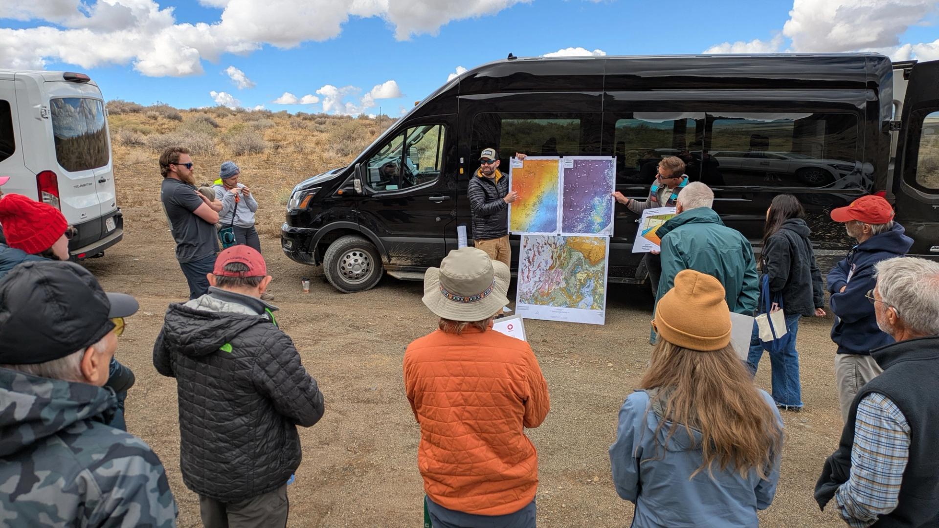 Field trip participants surrounding a speaker with a poster of three geologic maps in front of a black sprinter van.