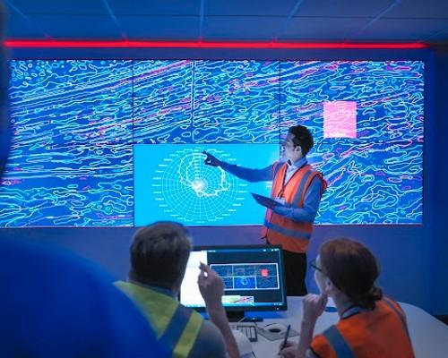 A person in a safety vest points to a large, illuminated screen displaying complex blue patterns. Two seated people observe, suggesting a data analysis setting.