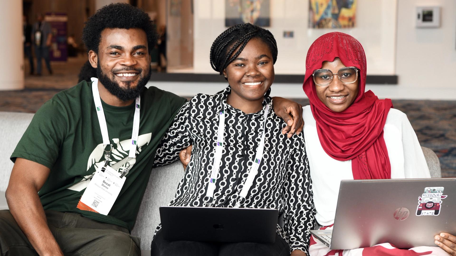Three people sit together on a couch, smiling. Two hold laptops, one wears a red headscarf. They appear cheerful and engaged in a bright room.