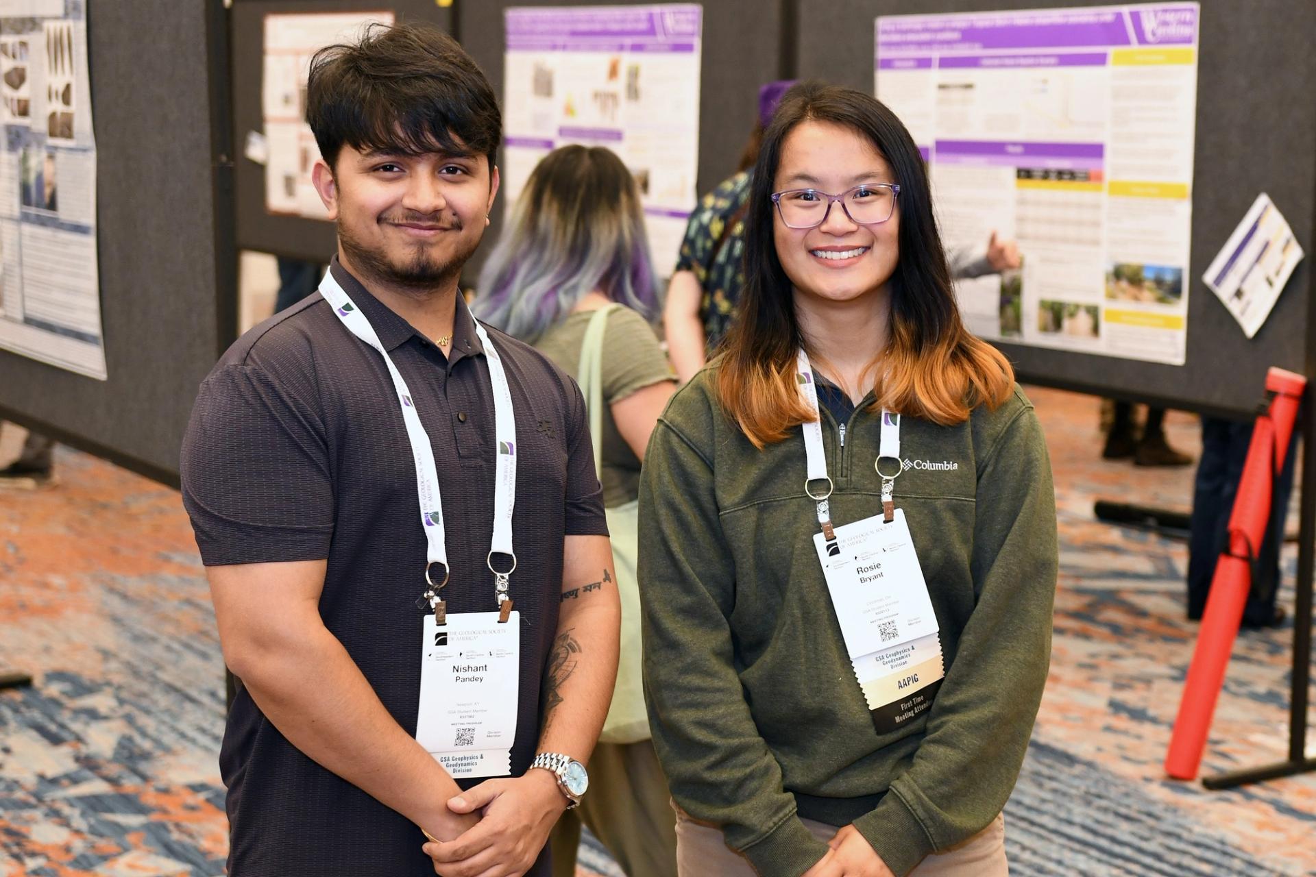 Two people smiling at a conference poster session, wearing name tags. They stand in front of display boards with research posters. The atmosphere is professional and friendly.