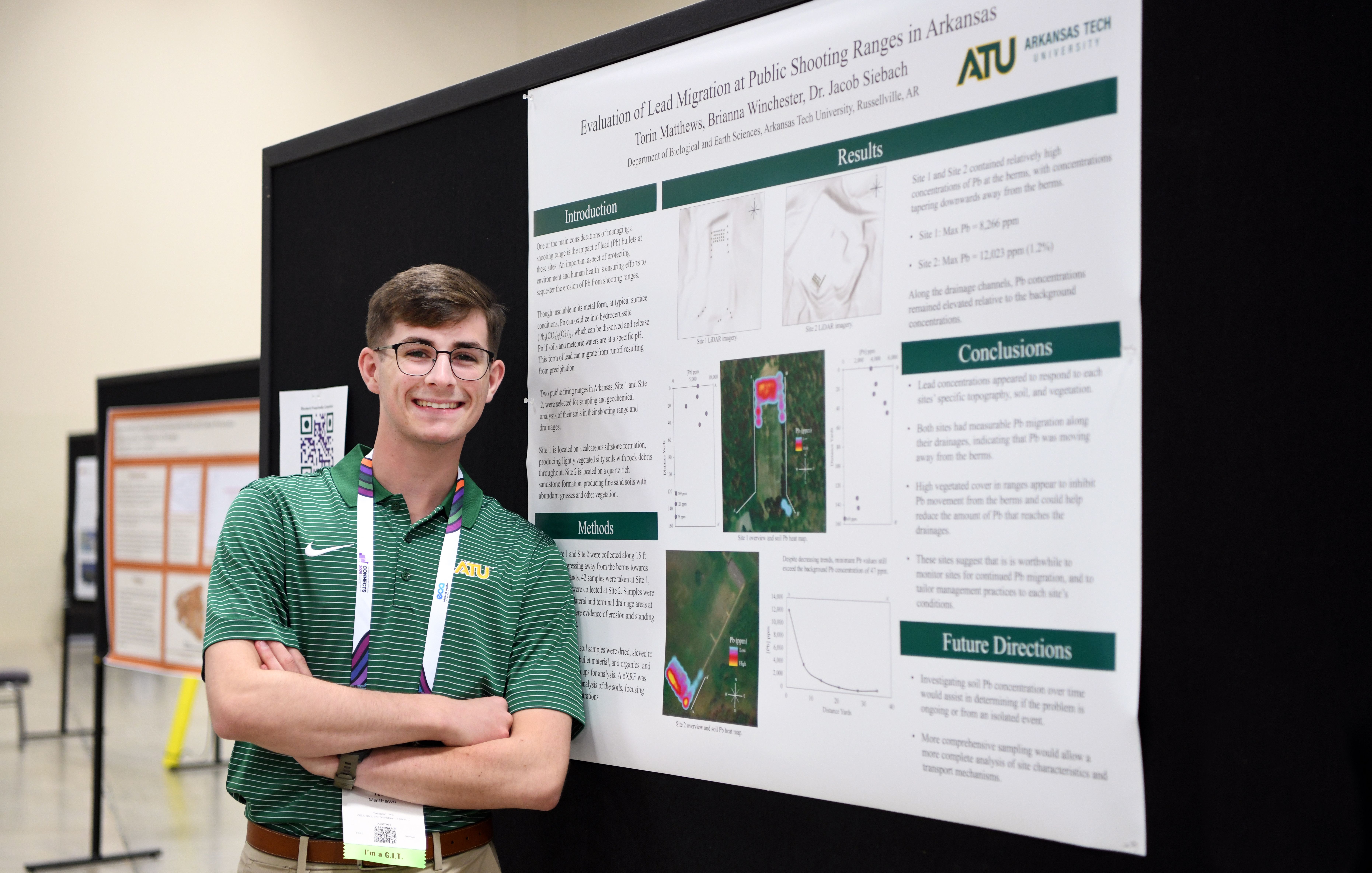 A smiling young male GSA conference attendee wearing an Arkansas Tech University polo shirt and conference badge stands with arms crossed in front of his research poster titled 'Evaluation of Lead Migration at Public Shooting Ranges in Arkansas' during a poster session.