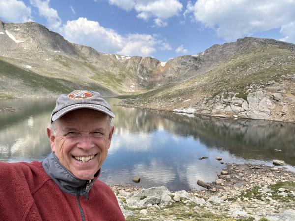 A man in a red and great sweatshirt and gray basebal cap smiles in front of a lake surrounded by mountains