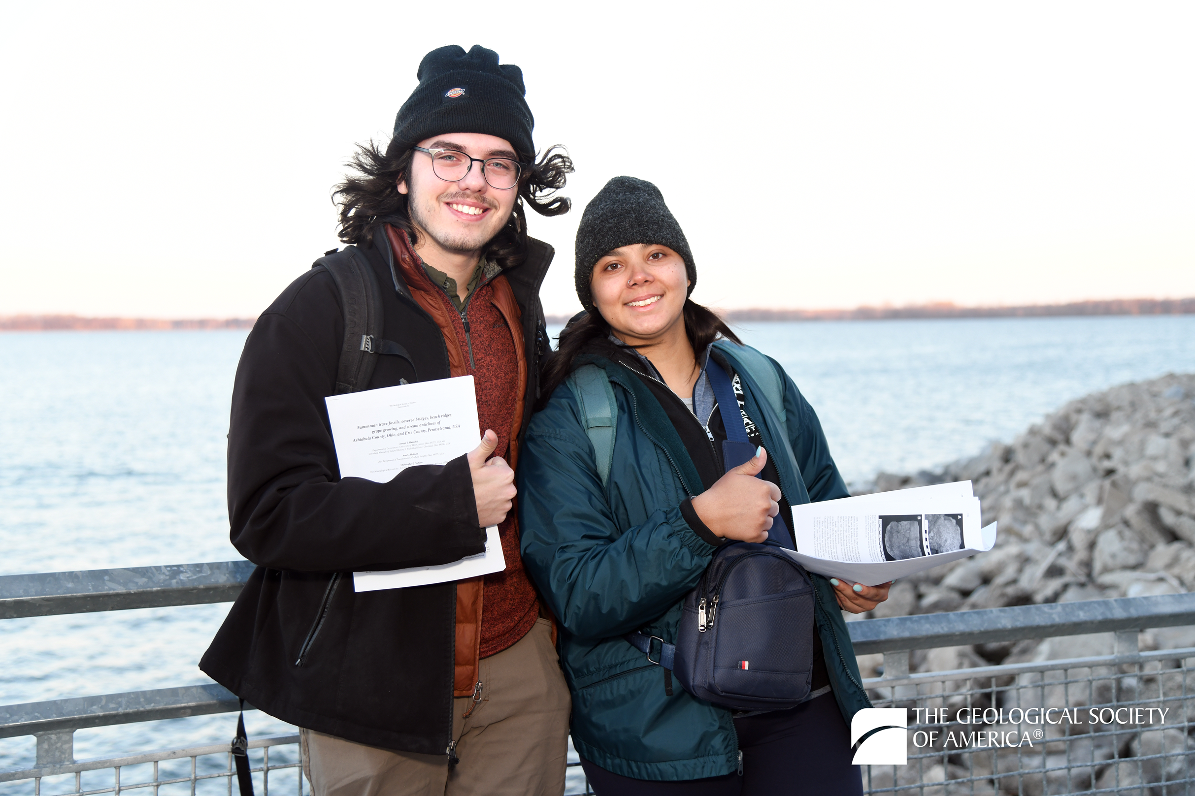 Two people in winter clothing stand on a pier by the water, smiling and giving thumbs up. They hold papers, suggesting an outdoor academic activity.