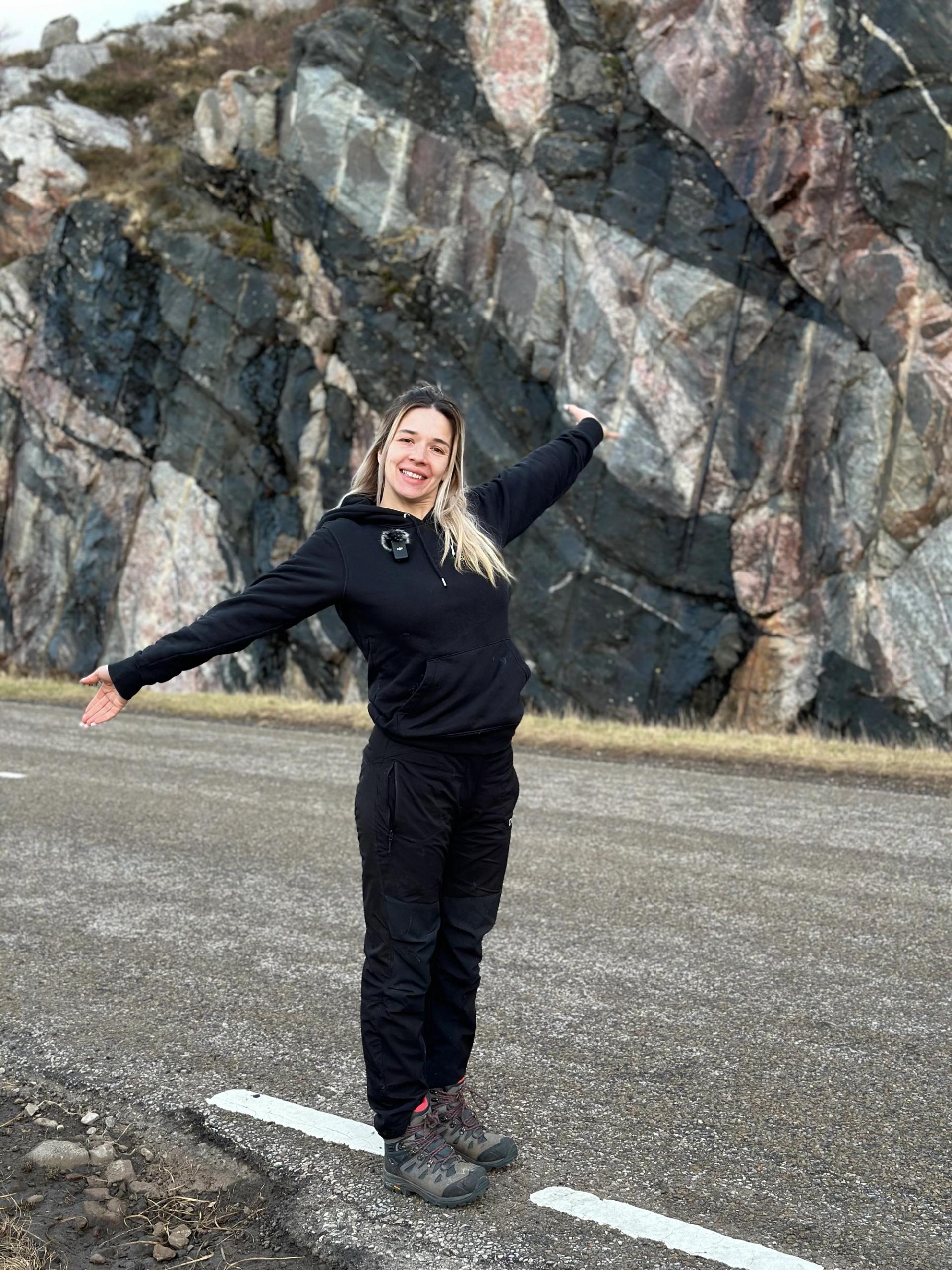 Luisa in front of a rock formation in Scotland standing with her arms in a T position wearing all black and smiling.