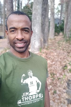 Mark Little smiles, wearing an olive green Jim Thorpe Fitness Center t-shirt, poses for an outdoor portrait in a wooded setting with trees and fallen leaves visible in the background.