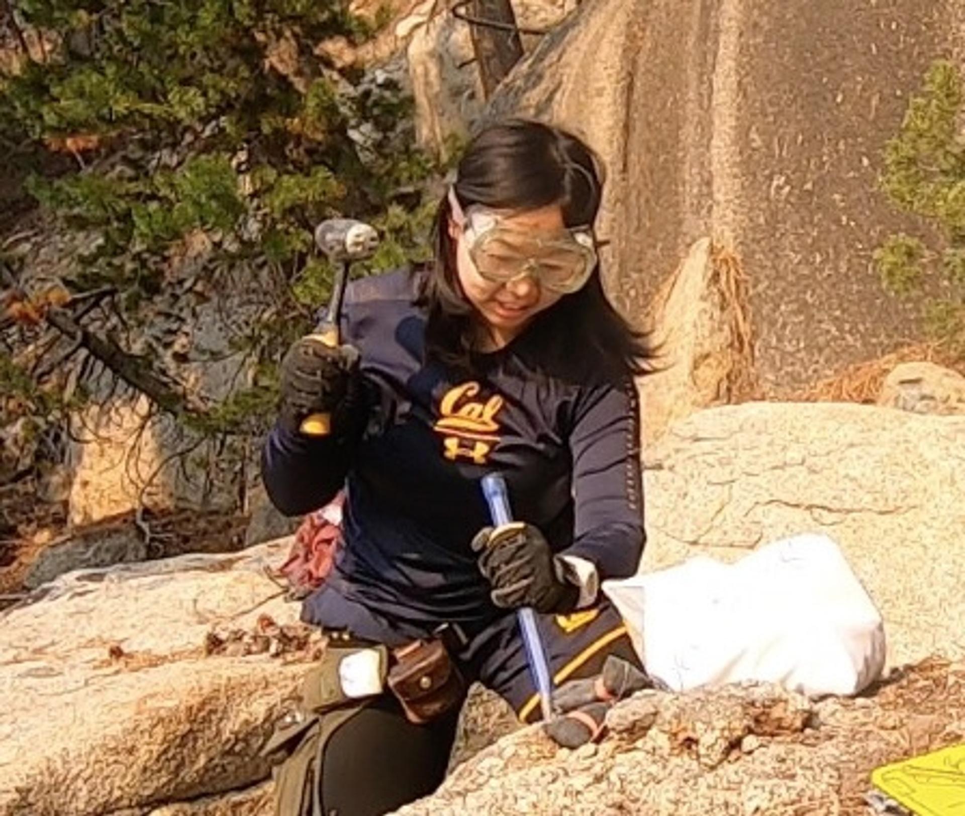 A female geoscientist, Yueyi Che, wearing safety goggles, gloves, and a UC Berkeley shirt, uses a rock hammer and chisel to collect samples from a granite rock outcrop during a field research expedition, with pine trees and rocky terrain visible in the background.
