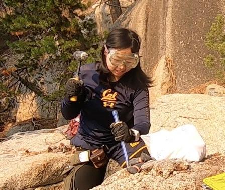 A female geoscientist, Yueyi Che, wearing safety goggles, gloves, and a UC Berkeley shirt, uses a rock hammer and chisel to collect samples from a granite rock outcrop during a field research expedition, with pine trees and rocky terrain visible in the background.