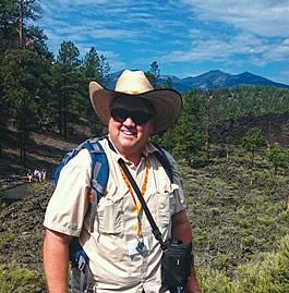 Steve Semken, smiling, wearing a wide-brimmed cowboy hat, sunglasses, and a field backpack stands on a dark volcanic lava landscape with pine trees and a mountain peak visible in the background.