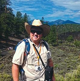 Steve Semken, smiling, wearing a wide-brimmed cowboy hat, sunglasses, and a field backpack stands on a dark volcanic lava landscape with pine trees and a mountain peak visible in the background.