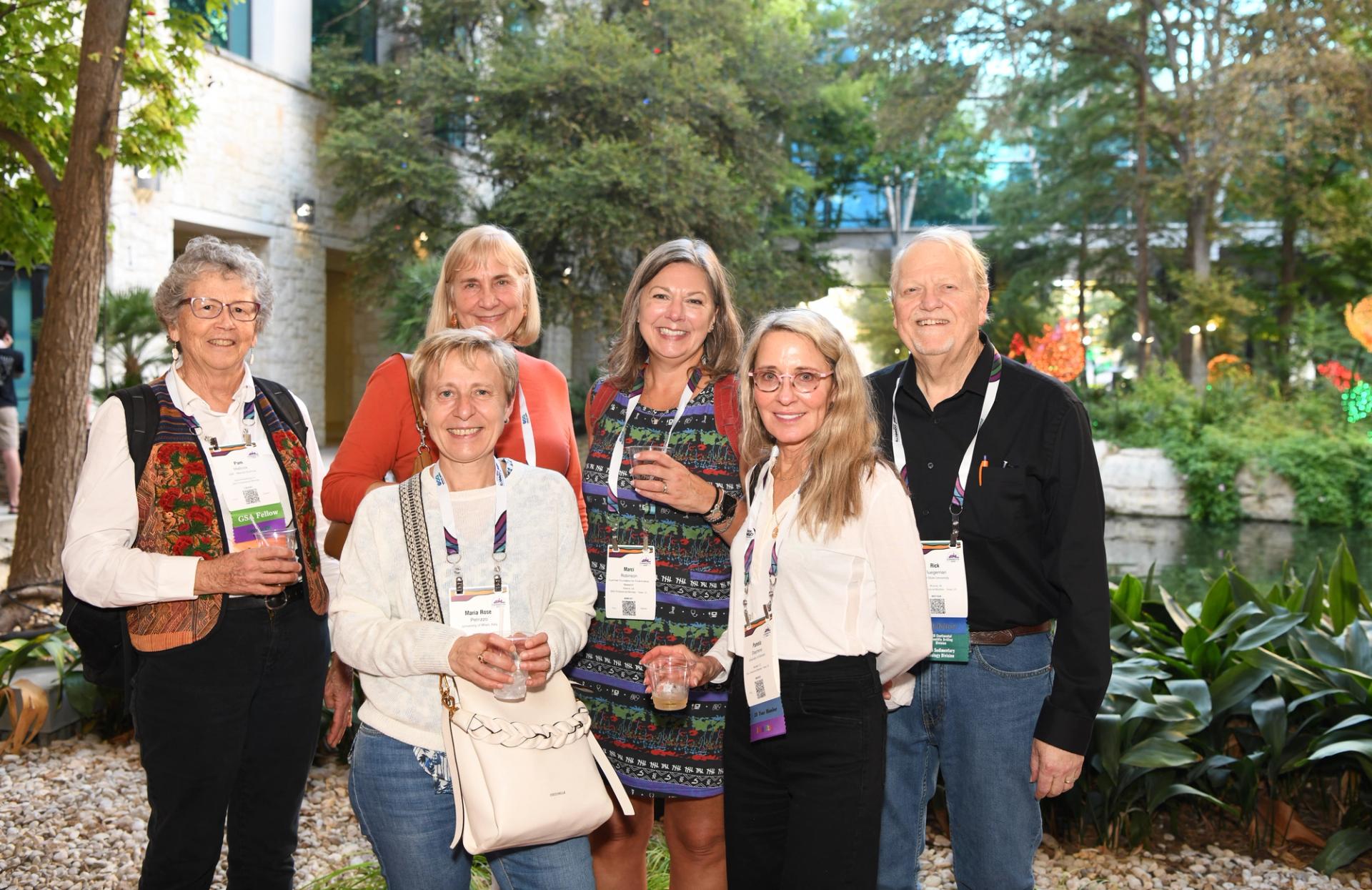 Six GSA Connects 2025 attendees wearing conference badges pose together outdoors during an evening icebreaker networking reception surrounded by trees, greenery, and a landscaped water feature in San Antonio, Texas, USA.