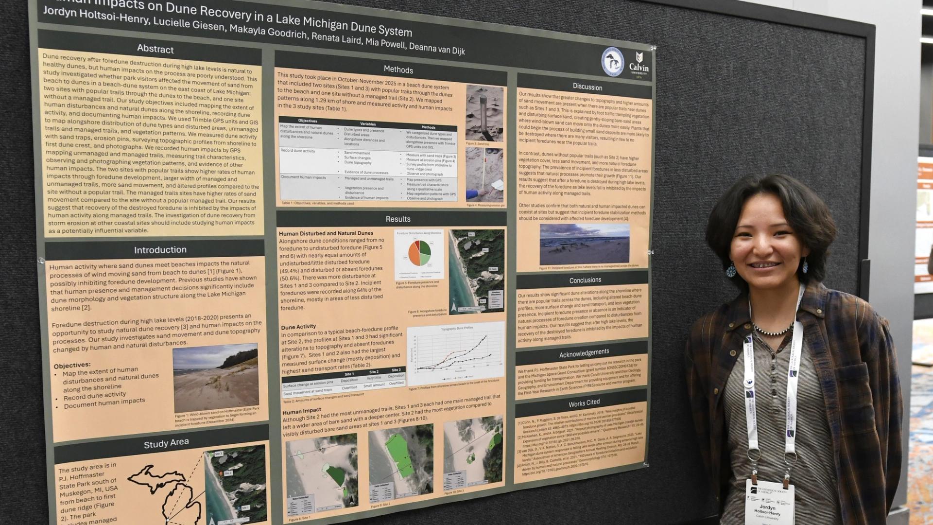 A person stands smiling next to a scientific poster titled "Human Impacts on Dune Recovery in a Lake Michigan Dune System," featuring text, charts, and images.