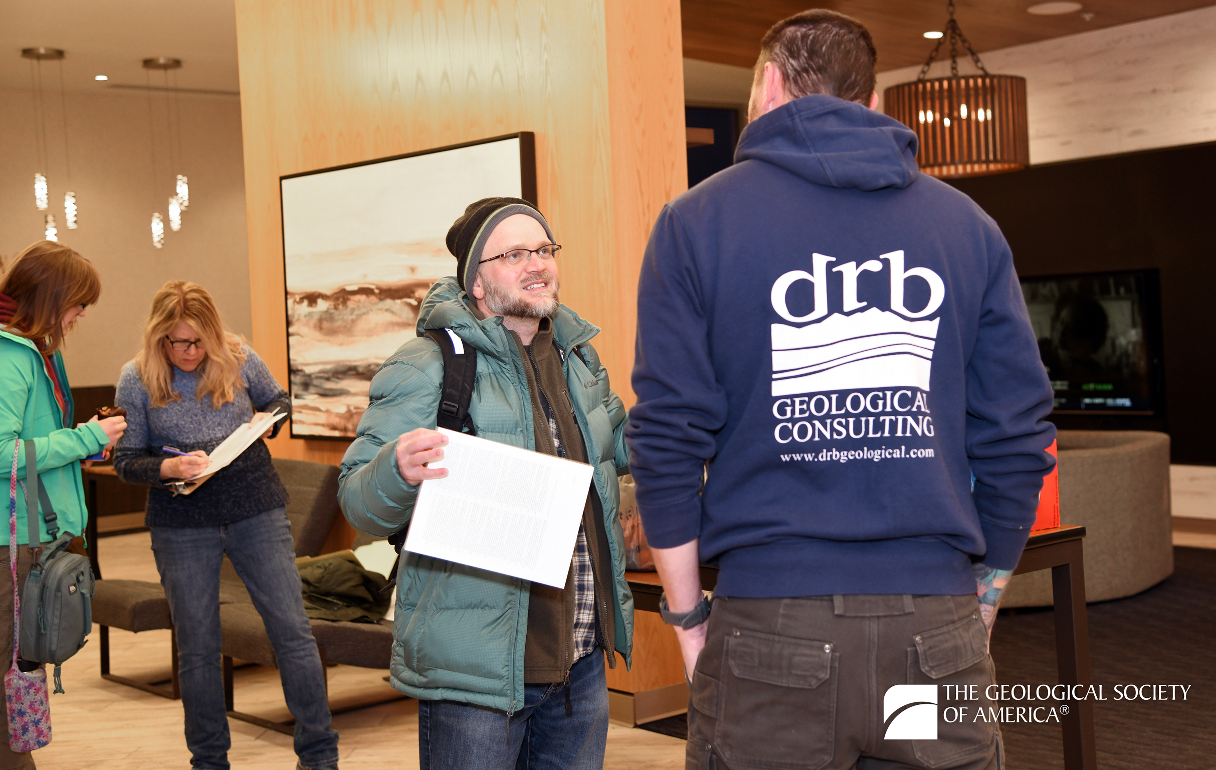 Two people converse in a modern lobby; one wears a "drb Geological Consulting" hoodie, while others in the background read documents. The mood is professional.