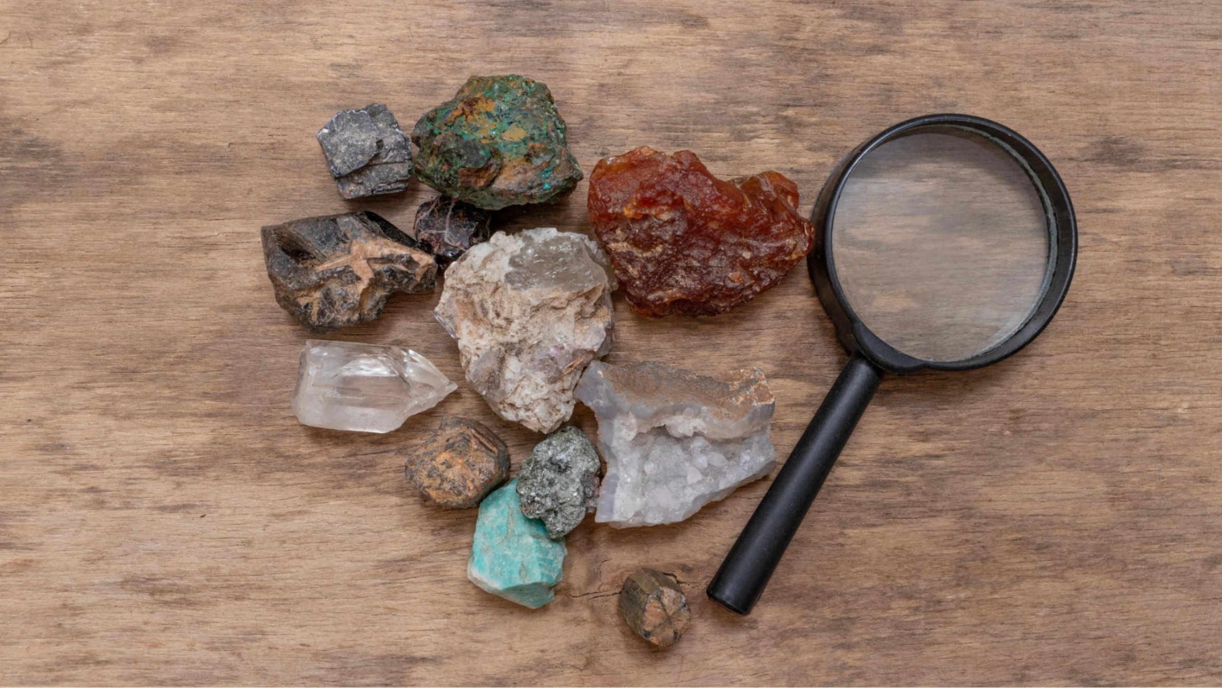 A collection of colorful minerals and stones lies on a wooden surface, with a black-handled magnifying glass beside them, suggesting exploration.