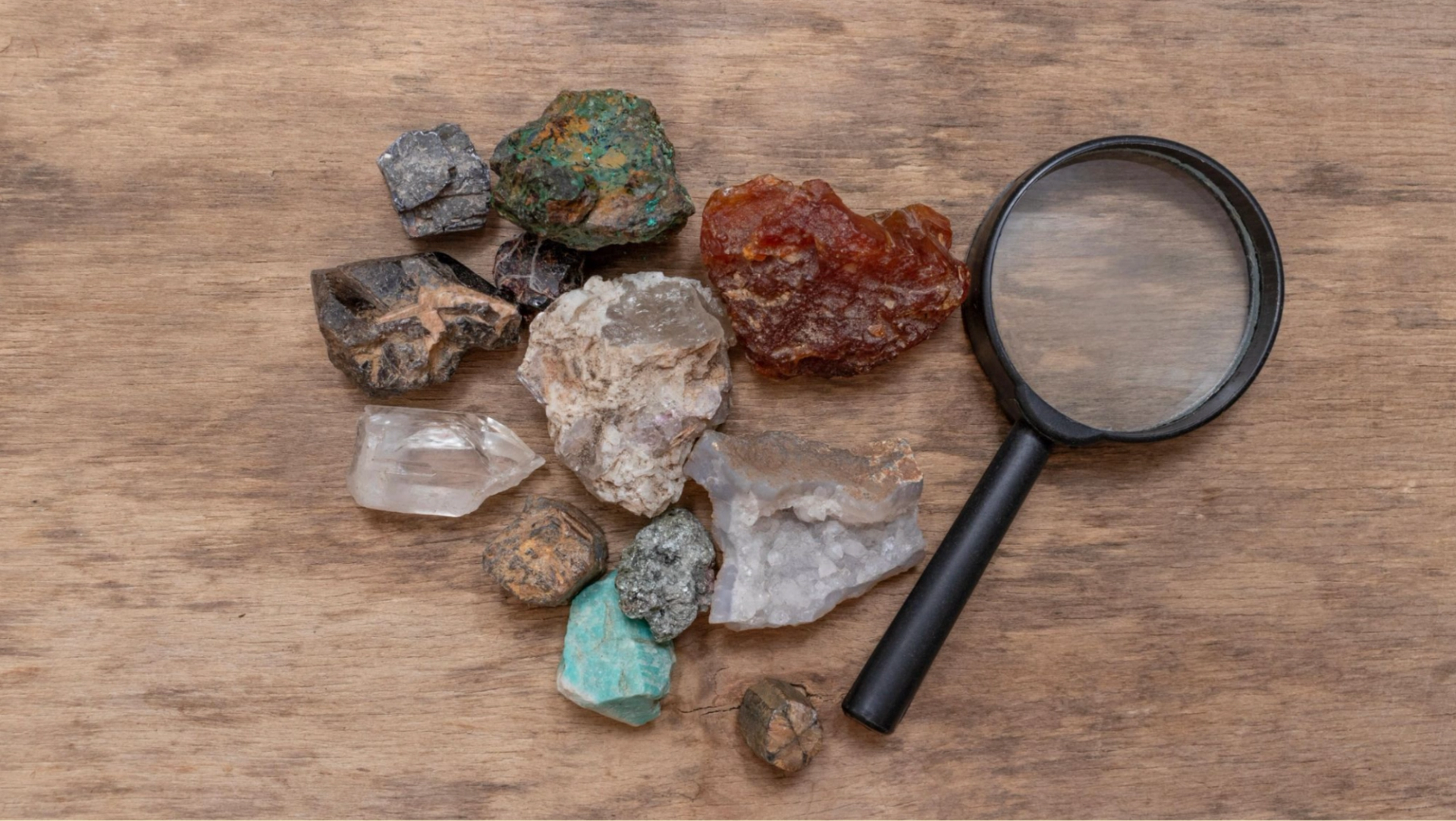 A collection of colorful minerals and stones lies on a wooden surface, with a black-handled magnifying glass beside them, suggesting exploration.