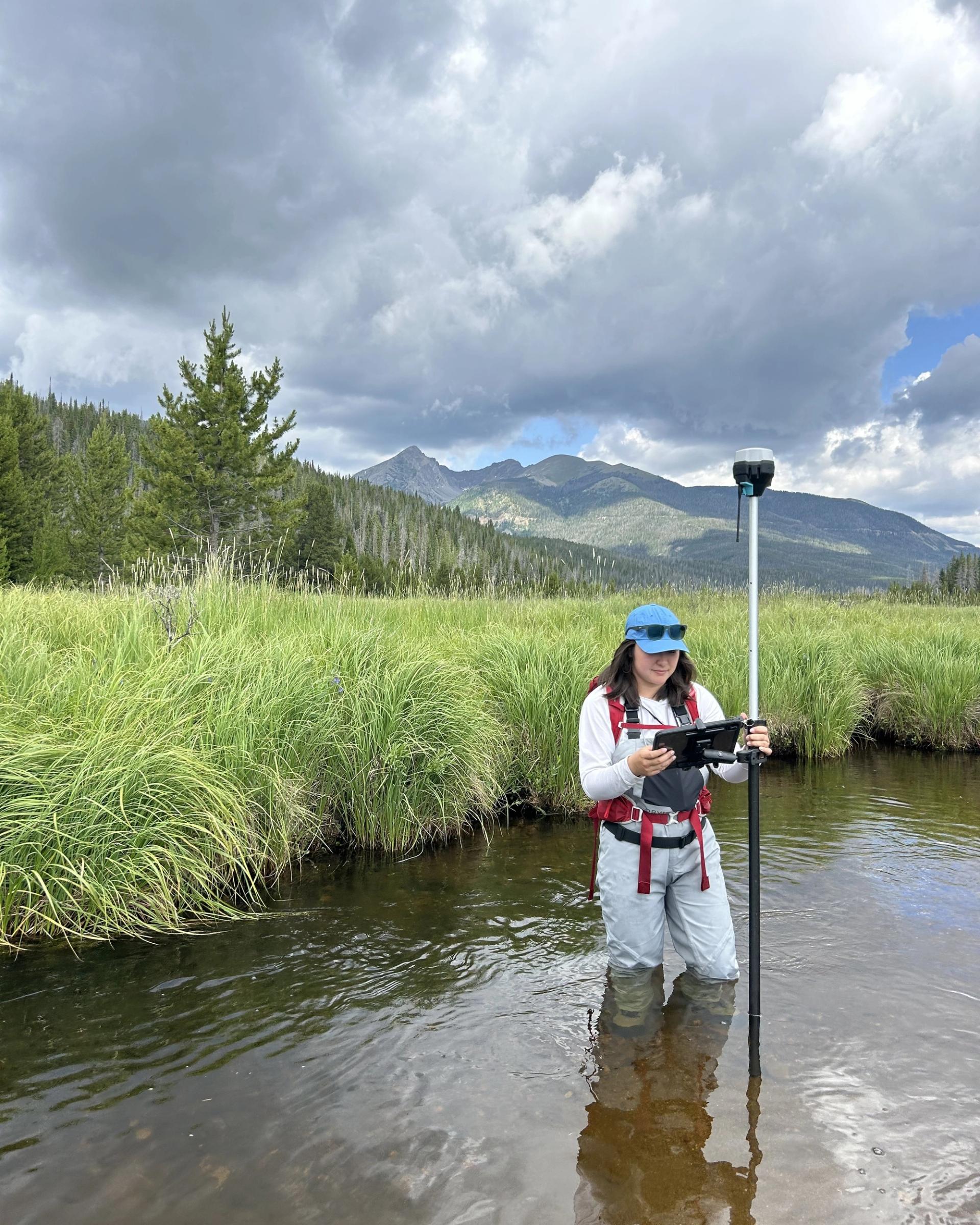 Photo of GSA Graduate Student Research Grant Recipient conducting fieldwork in a stream with mountains in the background