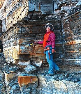 Kelsi Ustipak, a smiling female geoscientist, wears a red jacket, white helmet, and jeans as she stands against a dramatic towering wall of exposed layered sedimentary rock, pointing to a specific formation during a field research expedition.