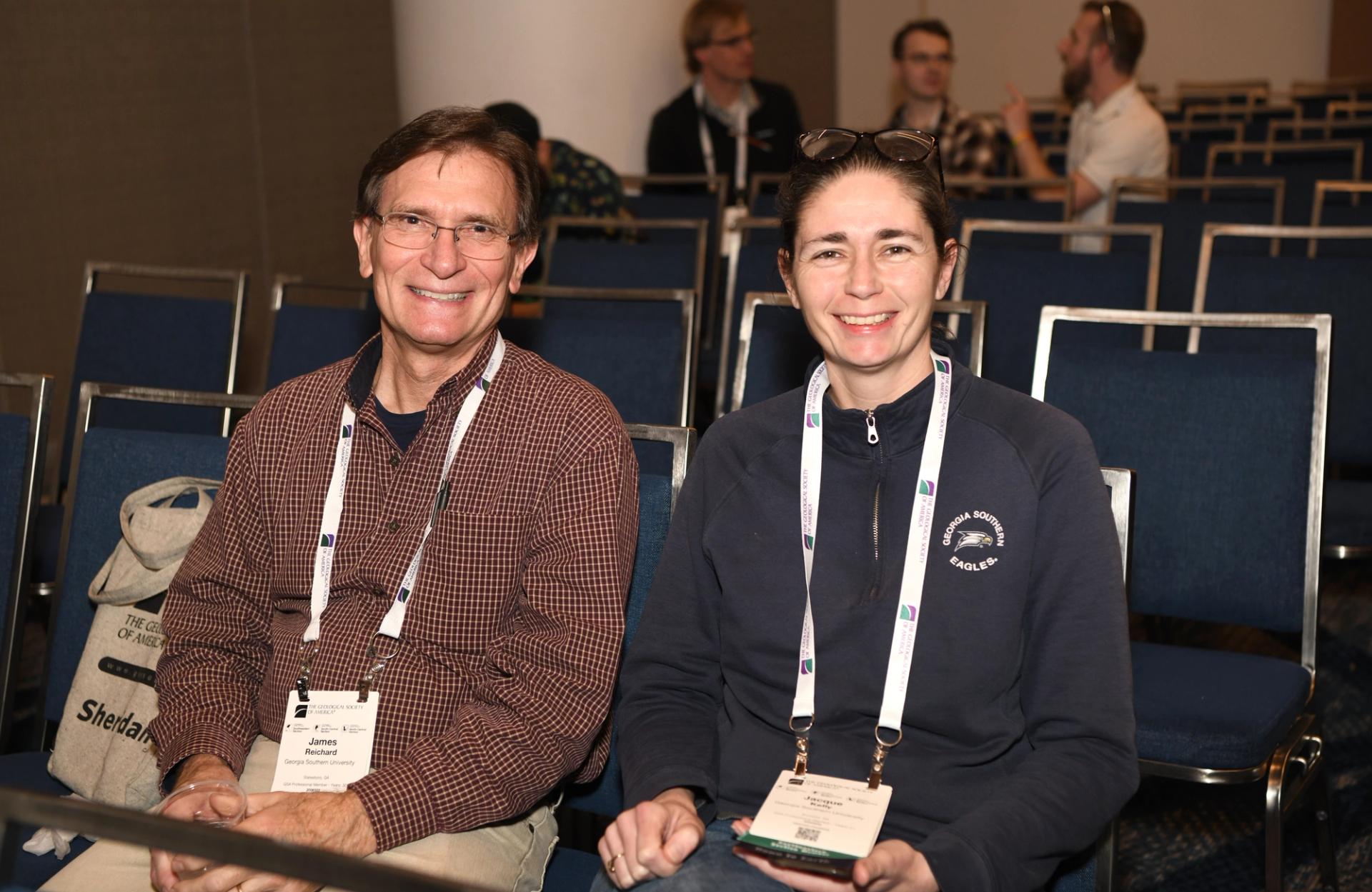Two smiling GSA Triple Joint Southeastern / North-Central / South-Central Section Meeting attendees wearing name badges and lanyards sit together in a blue-chaired session room, with other attendees visible in the background.