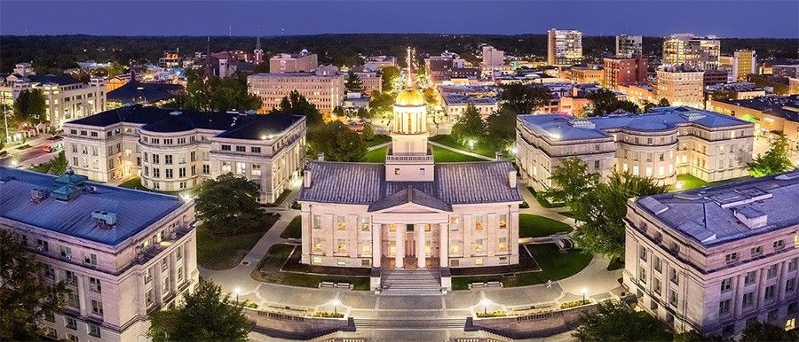 Aerial view of Iowa at dusk featuring a neoclassical building with a golden dome, surrounded by lit pathways and modern cityscape in the background.