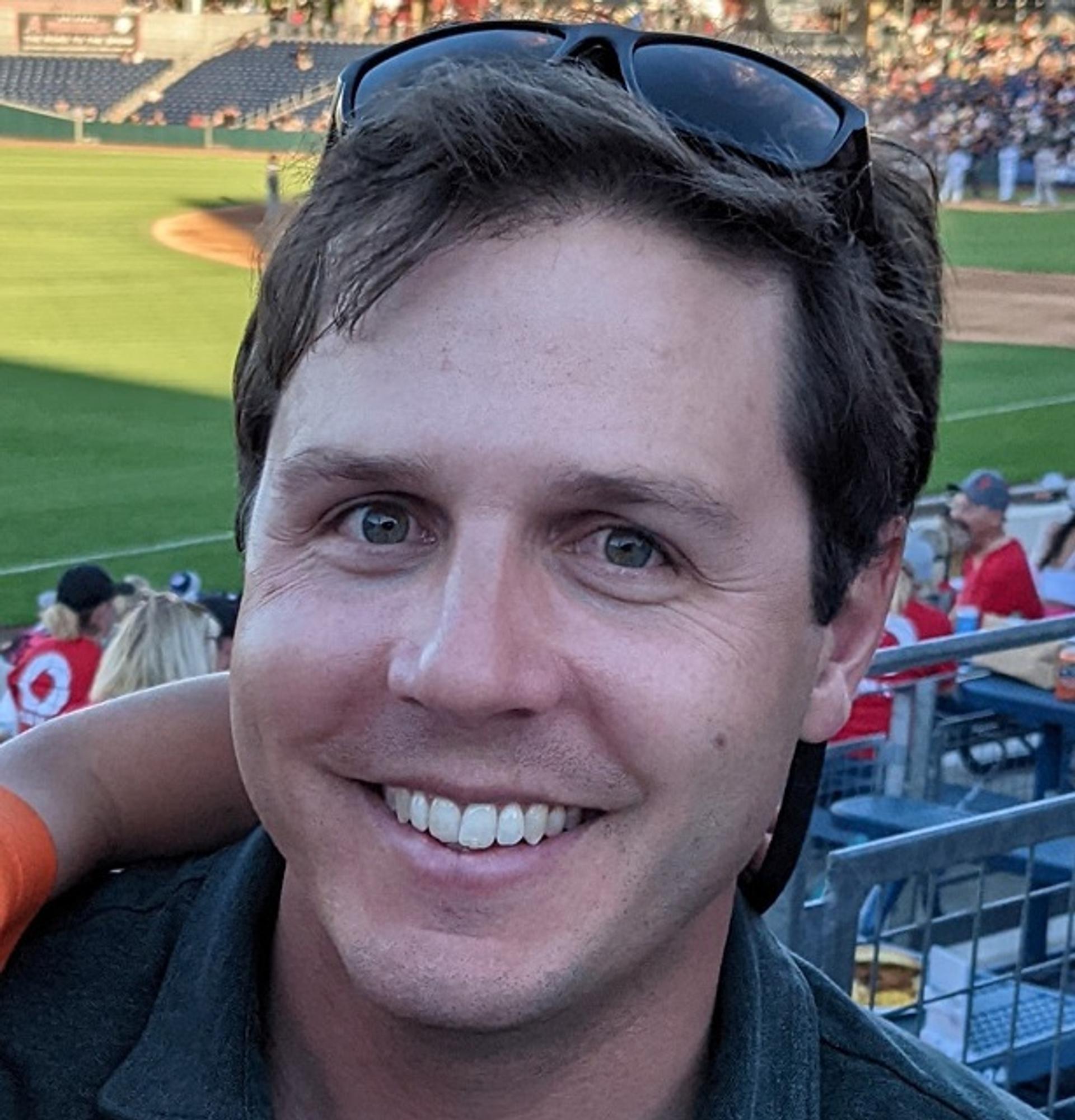 Andrew V. Zuza wearing a collared shirt with the grass of a sports field in the background.
